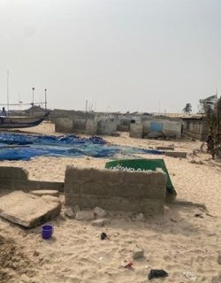 The image depicts a beach with boats and a boat. The setting is outdoors with a sandy beach and the sky in the background. Additionally, there is a mention of damage to dwellings from flooding and coastal erosion in Anyamam, with communities continuing to reside in these locations due to 'place attachment'.