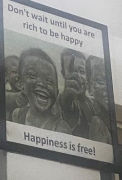 The image is a close-up of a poster with the following text: "Don't wait until you are rich to be happy. Happiness is free!" It includes the tags: text, human face, smile.
